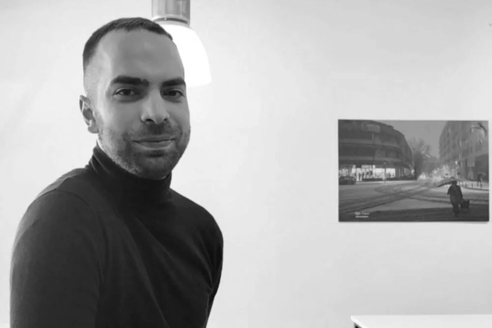 Marko Milačić u crnoj rolki, okrenut prema objektivu, u prostoriji sa uramljenom fotografijom na zidu.